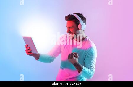 Winning online. Millennial guy in headphones celebrates win, holding tablet Stock Photo