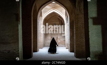 Isfahan, Iran - May 2019: Unidentified iranian woman in hijab black dress walking along the ancient columns of Jameh Mosque of Isfahan Stock Photo