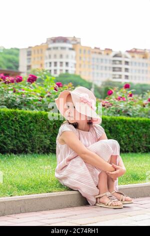 a beautiful little girl in a hat sits on the curb and looks away Stock Photo