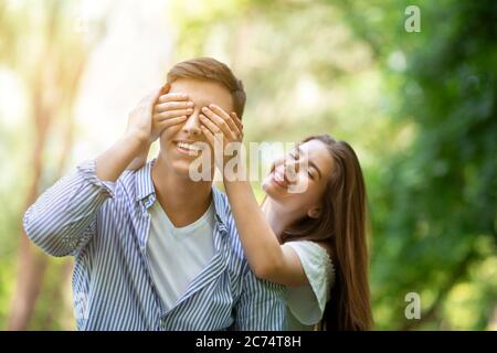 Happy together. Playful young girl closing her boyfriend's eyes at park, surprising him outdoors Stock Photo