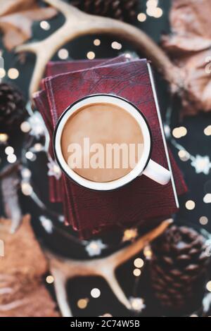 Hot, steaming cup of coffee over a stack of books with dried autumn leaves, deer antlers, pines cones, and sparkling lights. Selective focus on mug wi Stock Photo