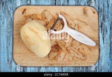 Raw potato peeled on a wooden chopping board with peeler cut out and isolated on a blue background Stock Photo