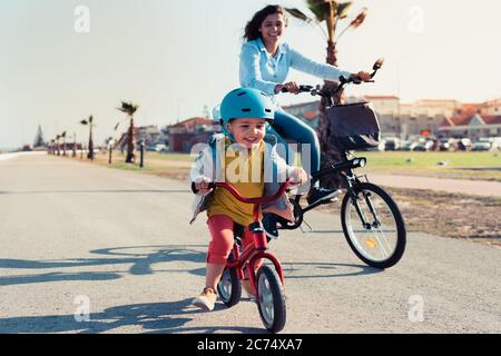 Little kid riding a balance bike with his mother on a bicycle in a city park Stock Photo