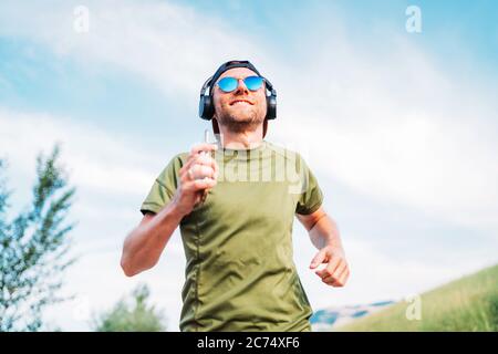Bearded man in a baseball cap, wireless headphones and blue sunglasses cheerful smiling running evening run and holding in hand modern smartphone device Stock Photo