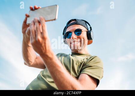 Bearded man in a baseball cap, wireless headphones, and blue sunglasses cheerful smiling taking a selfie picture using a modern smartphone device. Stock Photo
