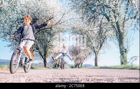 Father and son having fun spreading wide legs when riding bicycles on country road under blossom trees. Healthy sporty lifestyle concept image. Stock Photo