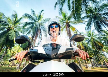 Happy smiling and screaming male tourist in helmet and sunglasses riding motorbike  scooter during his tropical vacation under palm trees Stock Photo - Alamy