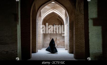 Isfahan, Iran - May 2019: Unidentified iranian woman in hijab black dress walking along the ancient columns of Jameh Mosque of Isfahan Stock Photo