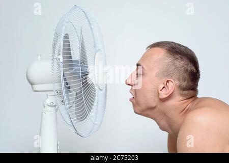 Man with stubble on his face suffers from the heat and trying to cool off near the fan. Stock Photo
