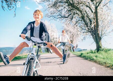 Father and son having fun spreading wide legs when riding bicycles on country road under blossom trees. Healthy sporty lifestyle concept image. Stock Photo