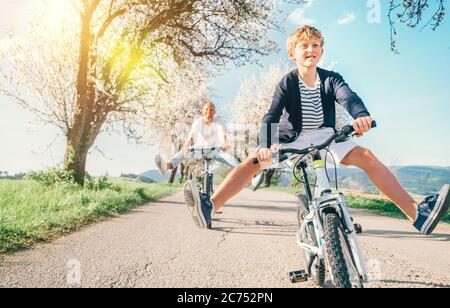 Father and son having fun spreading wide legs when riding bicycles on country road under blossom trees. Healthy sporty lifestyle concept image. Stock Photo