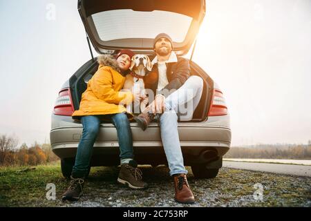 Father and son with beagle dog siting together in car trunk. Long auto journey break. Stock Photo