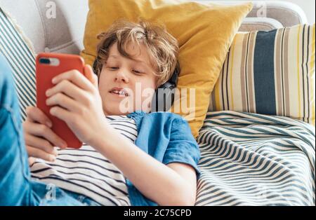 Preteen boy lying at home on cozy sofa dressed casual jeans and new sneakers listening to music and chatting using wireless headphones connected with Stock Photo
