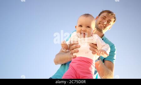 Father holds a small child in his arms on a background of a summer blue sky. Cute baby is smiling. Happy family outdoors. Right space for text. Stock Photo