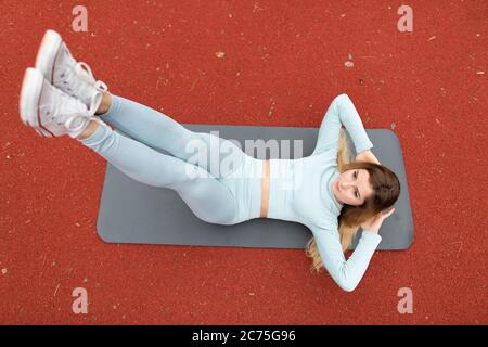 Young attractive sporty female trainer in gray clothes doing abs workout, performing leg raising and crunch exercise, lying on fitness mat outdoors on Stock Photo