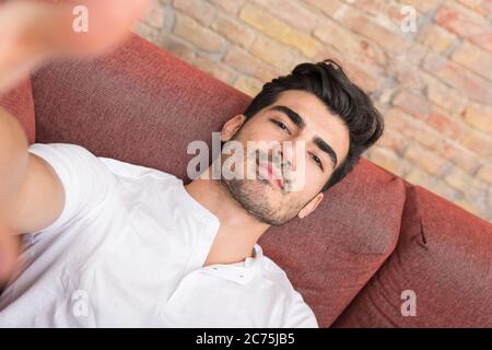 Portrait of a serious handsome young man talking a selfie while sitting on a sofa in a white tshirt from the perspective of the smartphone. Stock Photo