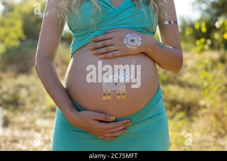 Tummy of pregnant blond girl with temporary silver golden tatoos, elephant symbol, expecting for baby boy, in blue outfit, at nature background Stock Photo