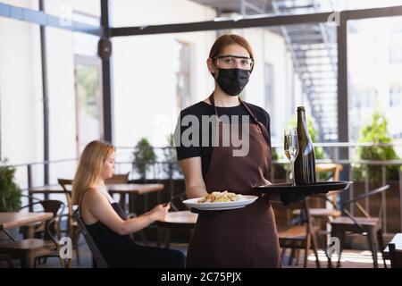 The waitress works in a restaurant in a medical mask, gloves during coronavirus pandemic. Representing new normal of service and safety. Putting the order, meals, drinks,. taking care of clients. Stock Photo