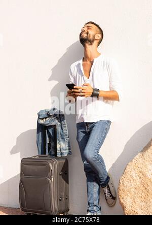 Happy young stylish caucasian man traveler smiling using the smartphone leaning against a white wall with traveling bag on the ground - Alwais connect Stock Photo