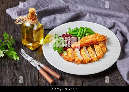 Chicken schnitzel served with beetroot salad and steamed crispy green bean on a white plate on a dark wooden table, horizontal view from above Stock Photo