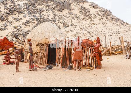 Himba tribe people indigenous to the Kunene Region of northern Namibia, south-west Africa gather round a typical mud hut in the village compound Stock Photo