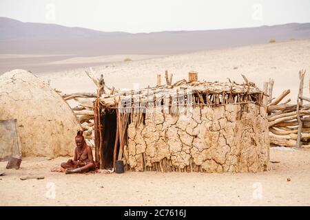 An old woman of the Himba tribe people indigenous to the Kunene Region of north Namibia, SW Africa sits outside a mud hut in the village compound Stock Photo