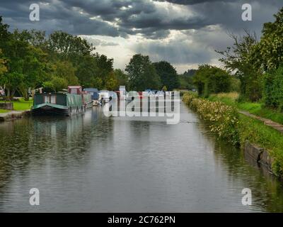 A row of canal narrowboats moored at permanent moorings in Halsall in Lancashire, taken on a grey, rainy day in summer.