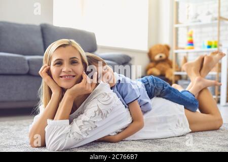 Beautiful young mother and her son lying on soft carpet at home. Cute boy hugging his mom on floor in living room