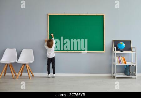 Back view of little boy writing or drawing something on chalkboard at classroom during lesson. Child studying at school Stock Photo