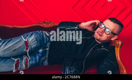 Handsome young man with glasses resting with smartphone on small sofa against red wall. Adult charming male talking on mobile phone, lying on his side Stock Photo