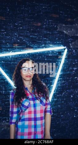 Young woman with glasses in colored plaid shirt posing on background shabby brick wall with glowing lamps. Portrait of charming female against Stock Photo