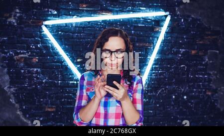 Young beautiful woman with glasses stands with mobile phone in hands on background brick wall with glowing lamps. Portrait of charming female flipping Stock Photo