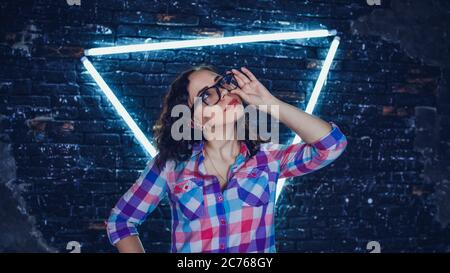 Young woman with glasses in colored plaid shirt posing on background shabby brick wall with glowing lamps. Portrait of charming female holding Stock Photo