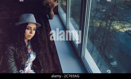 Young beautiful woman in hat and leather jacket sitting by windows. Portrait of pretty female looking out the window in daytime. Stock Photo