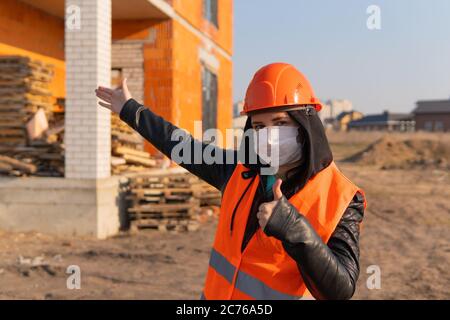 Female construction worker in overalls and in medical mask showing thumbs up and pointing at house under construction. Young woman in hard hat and Stock Photo