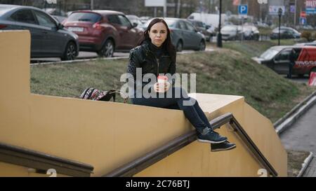 A young beautiful woman in casual clothes is sitting on a concrete fence and drinking a coffee on the background of the city. Stock Photo