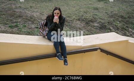 A young beautiful woman in casual clothes is sits on a concrete fence, looks away and holds a cup of coffee. Stock Photo