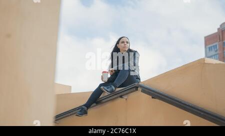 A young beautiful woman in casual clothes is sits on a concrete fence, looks away and holds a cup of coffee. Stock Photo