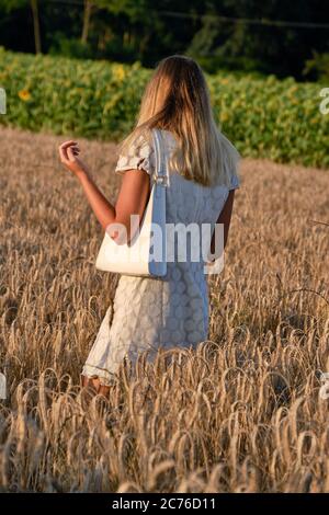 Full length shot of attractive young woman in field with sun shining in background. Female fashion model outdoors wearing white dress in countryside Stock Photo