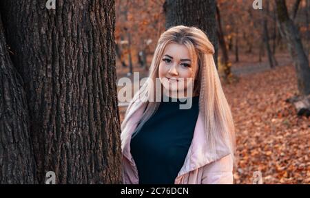 Beautiful woman looking out from behind tree. Pretty calm woman touching tree and looking at camera in quiet forest Stock Photo