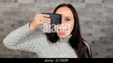 A young beautiful woman closes her eye with a phone. The woman is hiding behind a mobile phone. The concept of looking through the prism of the screen Stock Photo
