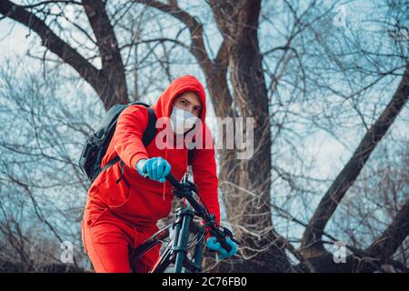 Young man in medical mask and gloves, ridding on bicycle in countryside. Male in red suit protecting yourself from diseases on walk. Concept of threat Stock Photo