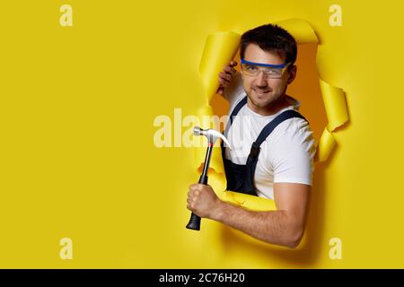 caucasian handyman worker in safety glasses and uniform holding hammer through hole in yellow paper. Stock Photo