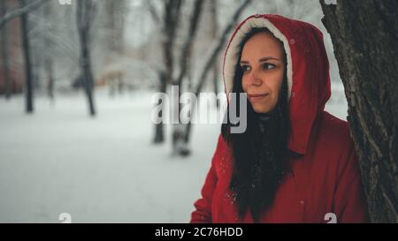 Portrait of young woman in red jacket and hood in winter season. Beautiful lady stands near tree and looks away. Fluffy snow envelops everything Stock Photo