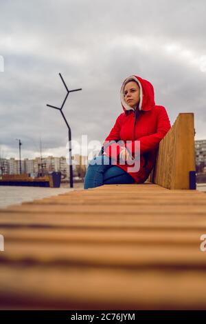 Young woman in red jacket and hood sitting on wooden bench in cloudy weather. Adult lady looking thoughtfully into distance. Stock Photo