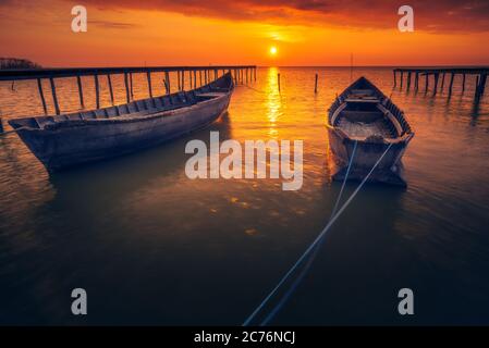 Two backlit fishing boats with the sun in the background at sunrise or sunset with some clouds on the sky and two wooden pontoons Stock Photo