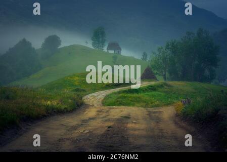 Two old abandoned barn houses on top of hills on a foggy dramatic day with a road leading to them and mountains in the background shot in Dumesti, Sal Stock Photo