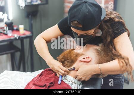 Barber woman shaves beard with a straight razor. Hairdresser equipment. Selective focus. Master in face mask Stock Photo
