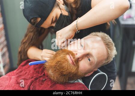 Barber woman shaves beard with a straight razor. Hairdresser equipment. Selective focus. Master in face mask Stock Photo