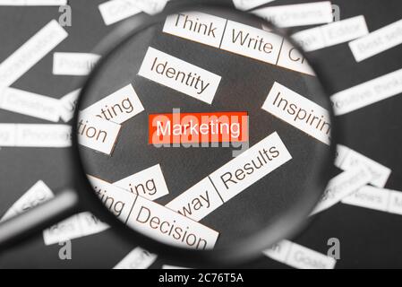 Magnifier glass over the red inscription marketing cut out of paper. Surrounded by other inscriptions on a dark background. Word cloud concept. Stock Photo